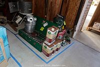 Wide angle photo showing the green Coleman 3-burner stove with pots and pans stacked on top, four fuel cans, and other camping equipment surrounding.
