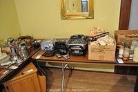 Wide view of table holding Farberware Electric Open Hearth Grill, multiple cameras, boxed glass cups, and assorted kitchen items.