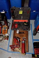 Lot with wooden creeper under black carrying case containing assorted tools, red jumper cables, and a red fire extinguisher arranged on a blue taped floor.