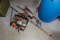 Photo of three fishing rods and a tackle box on the floor next to a large blue barrel.