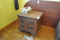 Front view of wooden side cabinet with two doors and stenciled panels, placed on floor next to wall. Surface has wear and some items on top.