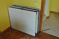 Two white rectangular folding tables stacked against wall, showing top surfaces and black trim.