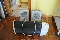 Three space heaters on wooden floor against yellow wall; one black and white Honeywell heater in front with mesh grille and two white Pelonis heaters behind, both with vents and handles.
