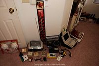 Photo showing assortment of vintage electronics, vacuum, clocks, and smoke alarms arranged on carpet against wall