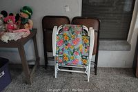 Six folding chairs including 4 brown padded folding chairs and 2 white metal outdoor folding chairs with floral canvas pattern stacked in front of brown chairs.