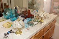 Photo of the lot showing two brass-colored lamps with white globes, blue textured glass bowls, brown textured glass goblet, blue ceramic swan and rooster figurines, glass storage dishes, floral trinket box, and decorative poodle figurine