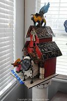 Front view of the red metal birdhouse with a cow sculpture and a yellow-black bird on top, displaying detailed embossed metal roofs and farm-themed art animals.