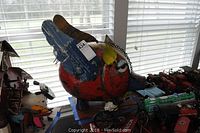 Pig head sculpture visible from left side, showing red body, blue ear, and some chain detail on the snout; background includes model airplanes and toys.