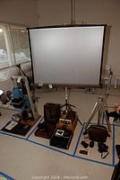 Wide view of vintage projection system setup showing Ansco Dualet projector, Kodak Movie Deck 475 projector, projection screen on tripod stand, aluminum tripod, leather case and binoculars on floor.
