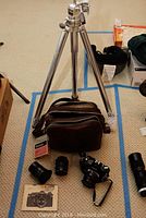 Photo showing the Canon EF 35mm film camera body with attached lens and cap, two additional lenses, tripod, camera bag, and filter box on carpeted floor.