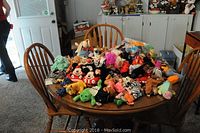 Wide view of a round wooden table covered with a large variety of Beanie Babies plush toys, including animals like elephants, zebras, frogs, and character toys like Mickey Mouse.