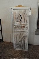 Front view of wooden outhouse-style wall shelf with moon cutout door and woven straw hat on top
