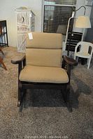Front view of wooden rocking chair with tan cushions showing seating and back padding, wood arms, and rocker base on carpeted floor.