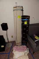 Three rolled rugs show two large rolled carpets and a small textured pink rug in front, all standing upright near a wall.