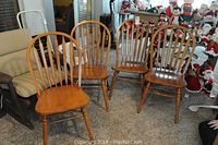 Four wooden spindle back chairs arranged in a semi-circle on carpeted floor. Chairs have a honey wood finish with vertical spindles on the backrest and turned legs.