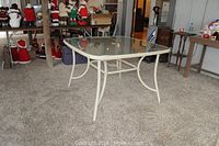 Photo of outdoor table showing entire table, curved white metal legs and frosted glass top in a well-lit room with other items in background.