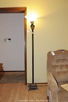 Full view of the tall floor lamp beside an armchair and wall thermostat, showing the black metal base, pole, and frosted glass shade lit.