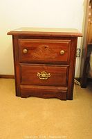 Front view of wooden bedside table with two drawers and metal hardware placed against wall on carpeted floor.