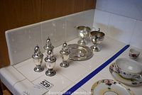 Overview of four silver shakers, round tray, creamer and sugar bowl arranged on tiled counter