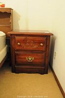 Wooden bedside table with two drawers. Top drawer has floral carvings and two round brass knobs, bottom drawer has ornate brass handles. Some wear visible.