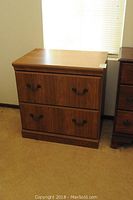 Closed view of wooden lateral file cabinet with two drawers and metal handles against a beige wall.