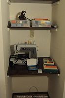 White Singer portable sewing machine with cords and two boxes of sewing thread and accessories on wooden shelves.