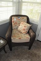 Front view of woven outdoor chair with floral cushions in a corner near windows showing cushion details and chair shape.