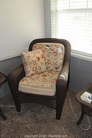 Brown wicker outdoor chair with floral-patterned seat and back cushions, plus throw pillow, placed indoors near windows.
