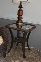 Full view of the square outdoor side table featuring a dark brown woven frame and glass top, shown with a bronze lamp on top and a carpeted floor background.