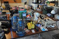 Overview of camping and utility items on table including multiple vintage lanterns, a boxed Coleman lantern, yellow flashlight, fuel bottles, tools, and a boom box.