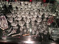 Overview of cut glass stemware including wine, sherbet, shot glasses and barware tools on black surface with mirror background.