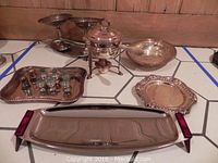 Overview of the lot showing two footed bowls, chafing dish with lid, silver plated trays, and salt and pepper shakers arranged on tiled floor