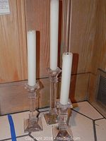 Three graduated Waterford crystal candlesticks standing on floor, each holding a white taper candle, showing their height differences and faceted clear crystal design.