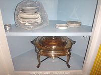 Photo showing stack of over 15 saucers and small plates partially wrapped in bubble wrap and a few small plates beside them on a shelf. Fire King silver plated casserole with glass lid and heating stand below on shelf.