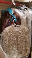 Light beige jacket with intricate gold and white embroidery hanging on a rack with other garments covered in plastic.