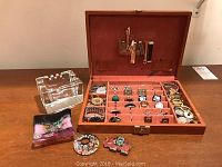 Open red suede-lined organizer box containing multiple cufflinks, tie clips, wristwatches, pins, buttons and fobs; clear square glass ashtray; glass dish and decorative pieces