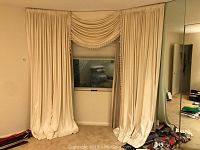 Ivory window drapes with two long panels and a swag valance trimmed with tassels, hung on a window with blinds behind them in a room with beige walls and carpet.
