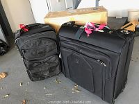 Two black luggage pieces side by side with original box behind them on floor. Rolling garment bag with pink ribbon on handle and carry on luggage with front pockets.