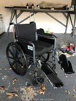 Full view of the black transport wheelchair showing seat, backrest, armrests, large rear wheels and foldable footrests with footplates.