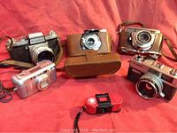 Overview of six vintage cameras including Argus 50 MM in leather case, Pratika, Kodak Retina II F, Canon Sure Shot digital, Canonnet QL17, and small red spy camera on red fabric background.