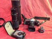 Shows full set of vintage Pentax Spotmatic camera with attached lens, telephoto zoom lens in case, Gossen Luna-Pro light meter open for viewing, metal tripod with light, and flash bulb on red fabric background.