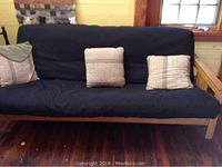 Full size futon with natural pine frame and navy mattress with three cushions placed on it, sitting on wooden floor below window.