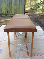 Fully set up brown massage table outdoors on cardboard with wooden legs and metal braces, vinyl top surface visible with rip