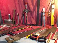 Wide shot showing all 27 pieces including saws, machetes, files, rulers, hand drill, spirit level, and hay bale hook on a table with red backdrop.