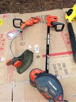 Photo showing two Black & Decker electric garden tools on cardboard: a string trimmer (weed wacker) with orange motor housing and flexible shaft, and an edger with Black & Decker logo on shaft and orange motor housing.