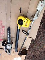 Full overview of all three tools on cardboard surface outside:  white Ryobi weed wacker, yellow Paramount leaf blower, and Craftsman chainsaw