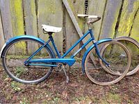 Side profile photo showing full blue step-through frame, white seat, original chain guard with 'Rodeo' label, rear and front wheel, rust on rims and frame, and an extra front wheel leaning beside bike.