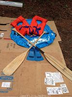 Photo showing all included water activity items: blue inflatable tube raft, two wooden paddles, two orange children's life jackets, and blue rubber flippers laid out on cardboard.