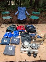 Photo showing a white two-burner gas stove with pots and pans, three camping chairs in green and blue fabric, three backpacks in red, green, and black, a beige backpack frame, two large waterproof bags and one small waterproof bag, and two blue 2-gallon water jugs.