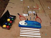 Full view of bocce balls in black metal case, badminton rackets, blue plastic badminton case and white extension poles laid on cardboard.